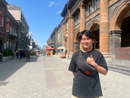 Yuki Kusakabe smiling on a pedestrian street lined with historic stone buildings in Gyumri