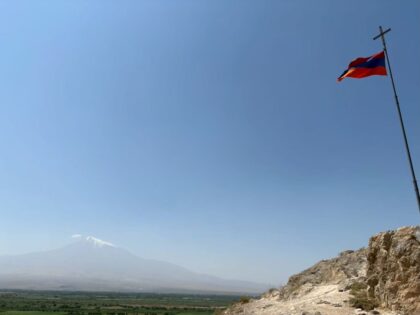 Snow-capped Mount Ararat in the distance, with the Armenian flag flying beneath a cross on a rocky hilltop
